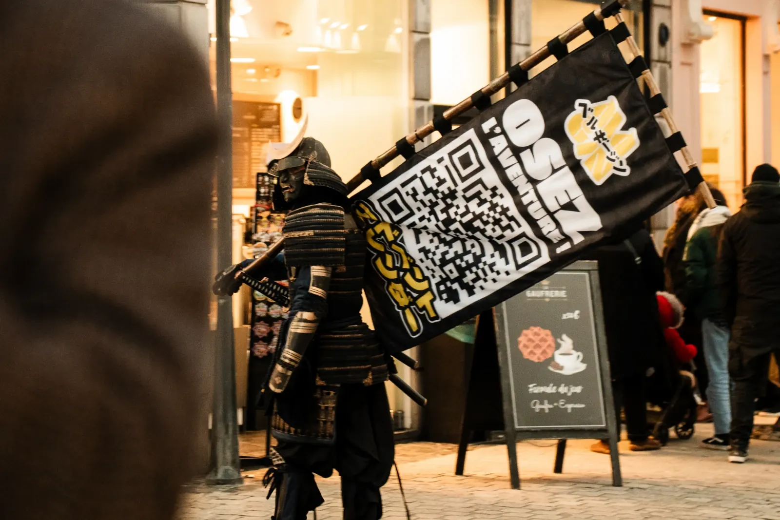Le samouraï MaGin et sa bannière « Osez l'aventure ! » dans une rue commerçante de Liège
